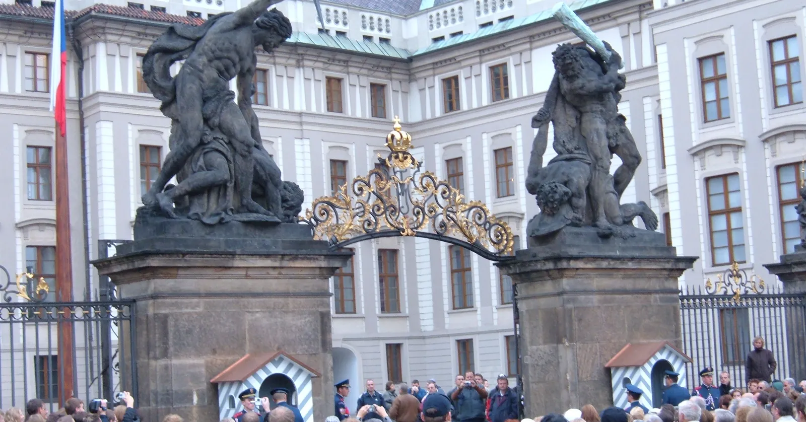 The main gate of Prague Castle, with the two Battling Titans statues on stone piers above a gilded wrought-iron gateway, sentry boxes beneath them and visitors gathered in front