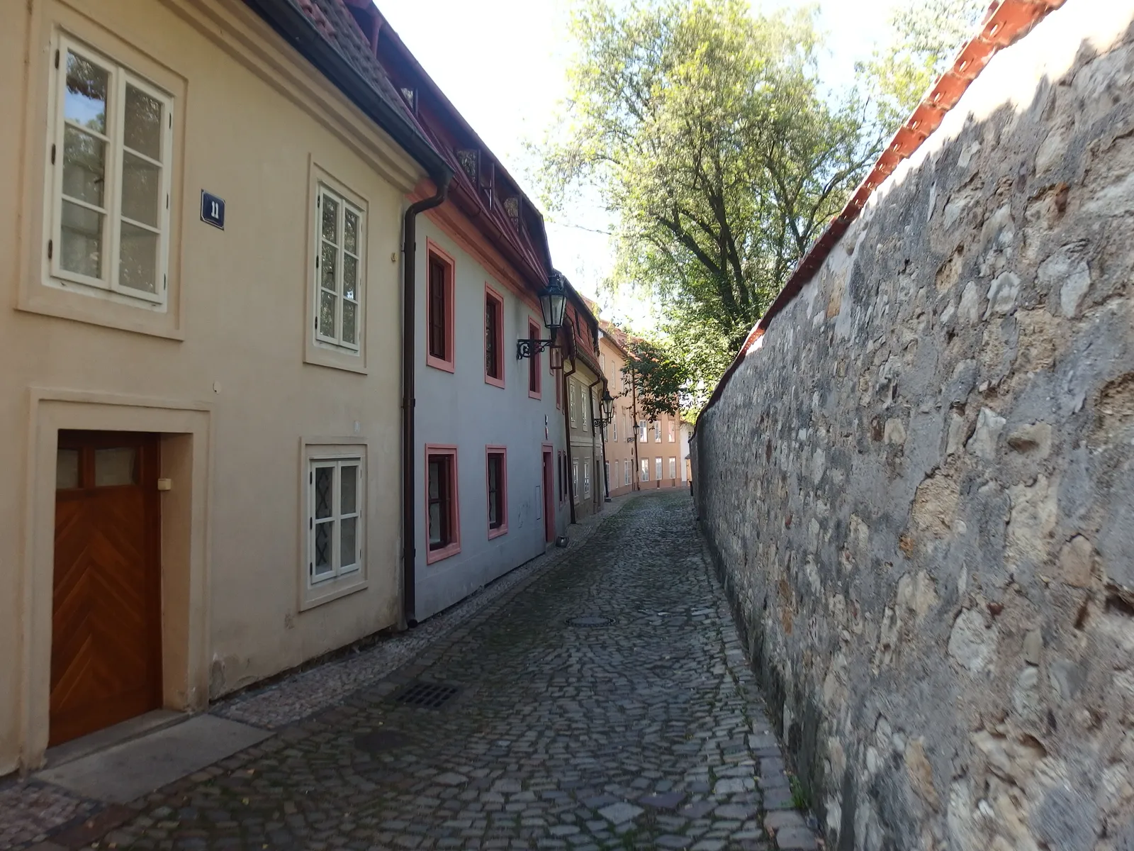 A narrow cobbled lane in Nový Svět behind Prague Castle, with small pastel houses and an iron lamp on one side and a high rough stone wall on the other, empty of people