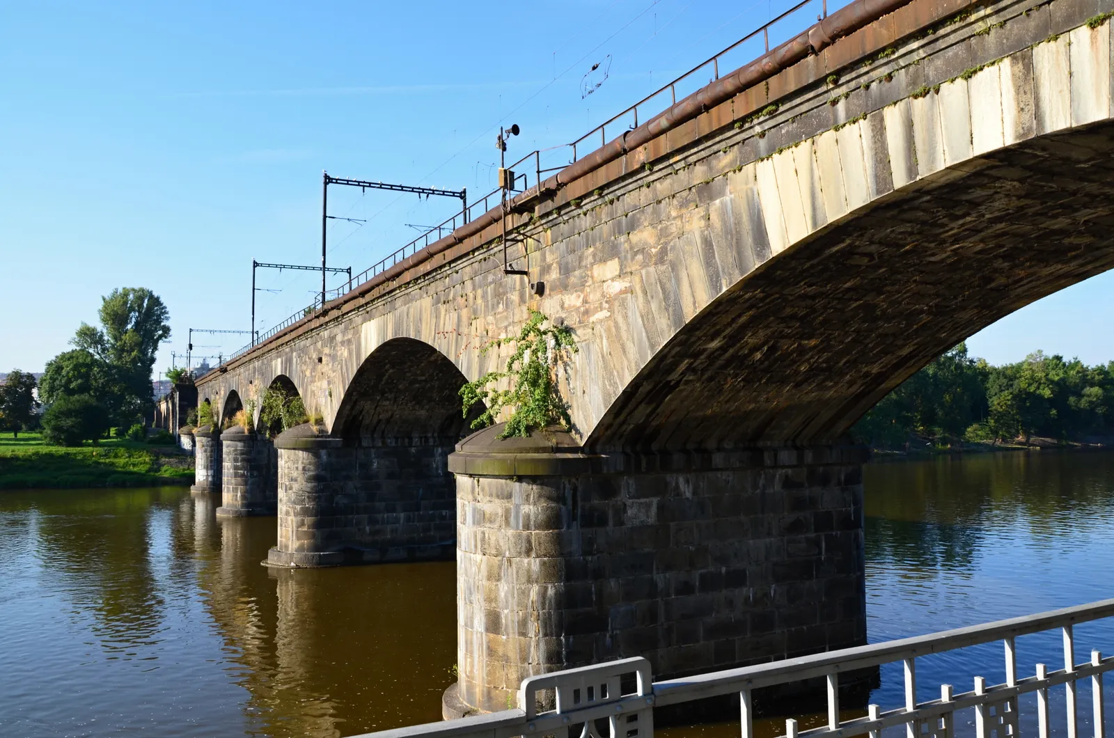 The Negrelli Viaduct in Prague, a long stone railway bridge on heavy masonry piers carrying track across the Vltava, seen from the embankment on a sunny day