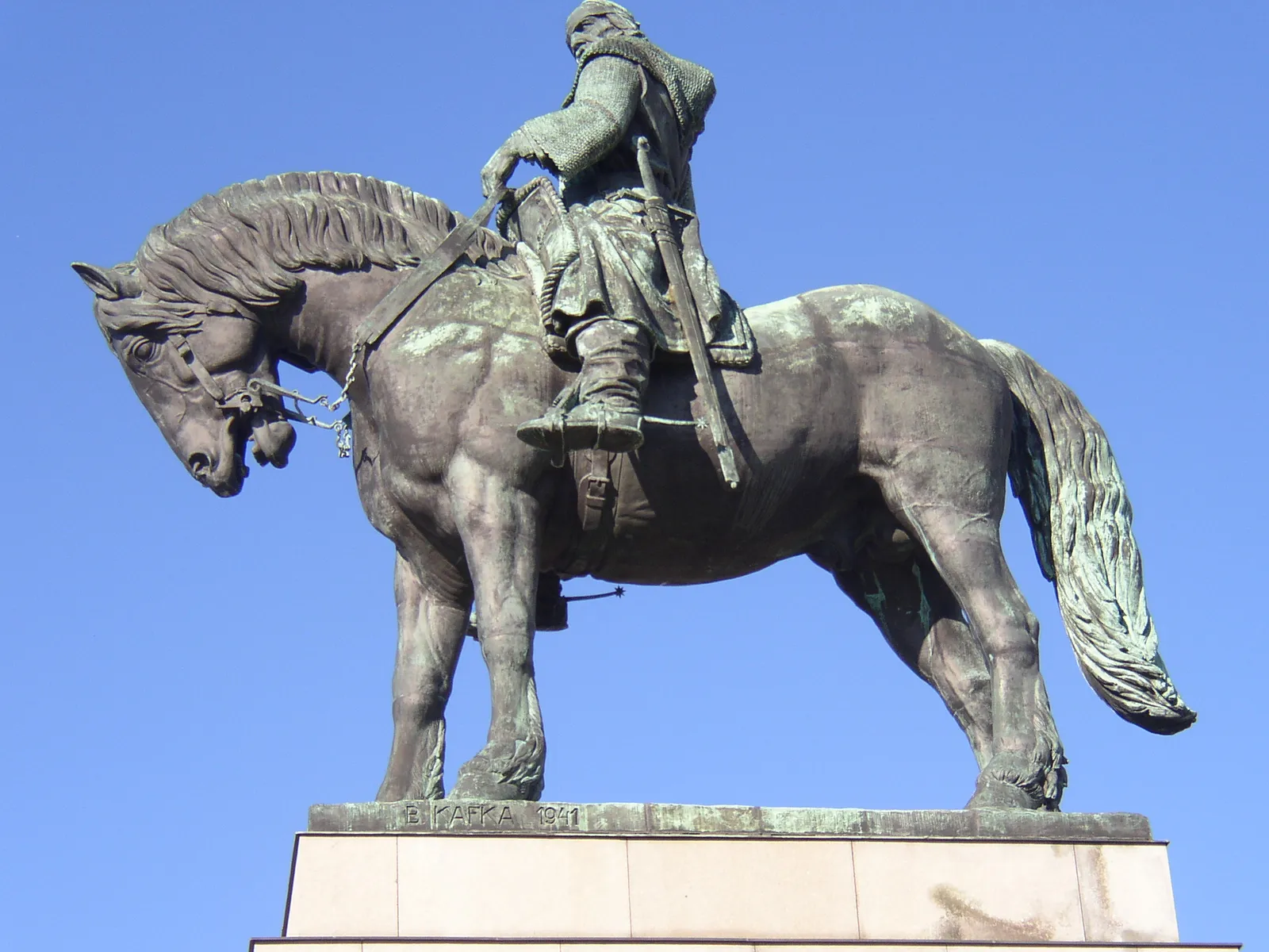 The bronze equestrian statue of Jan Žižka on Vítkov Hill seen side-on against a blue sky, the horse standing squarely on a stone plinth with the armoured rider holding a mace