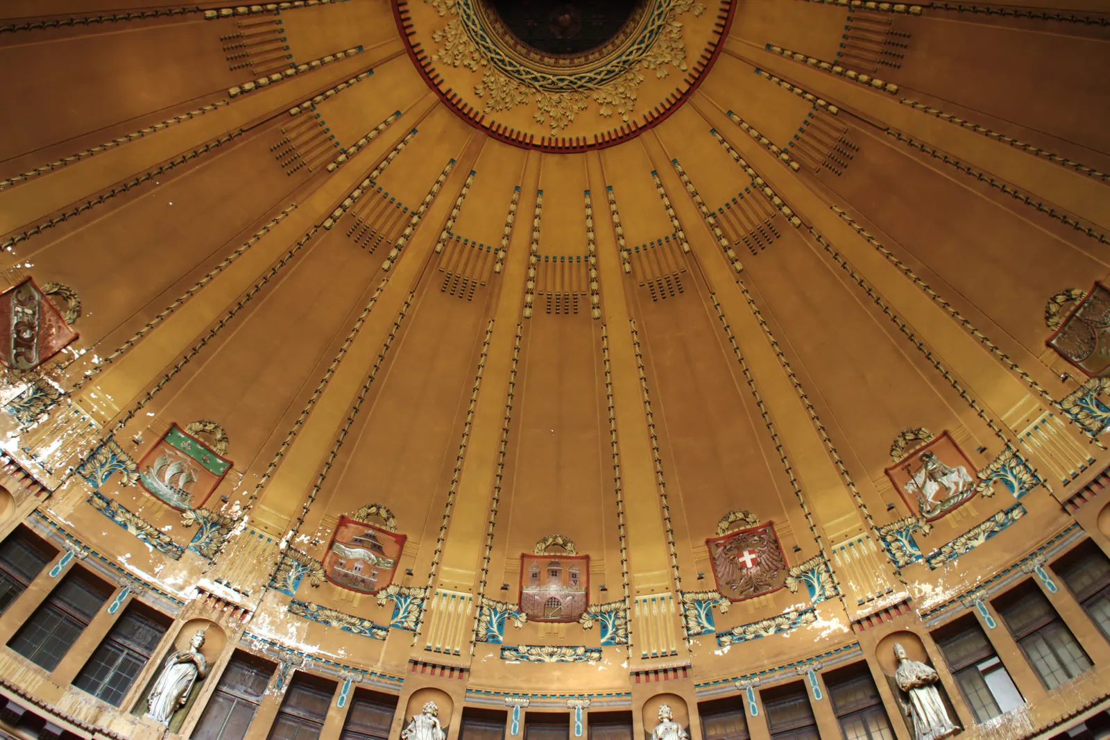 The ribbed ochre and gold dome inside the Art Nouveau hall of Prague main station, ringed with painted medallions bearing the coats of arms of Czech towns and with statues standing in niches below