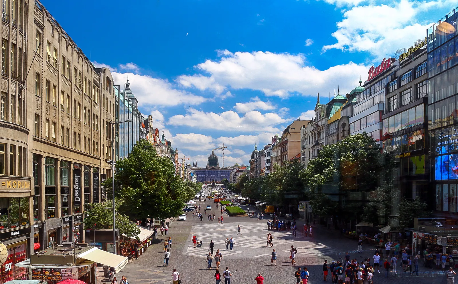Wenceslas Square in Prague looking uphill from the Můstek end on a sunny day, a wide boulevard lined with shops and trees, with the domed National Museum closing the view at the far end