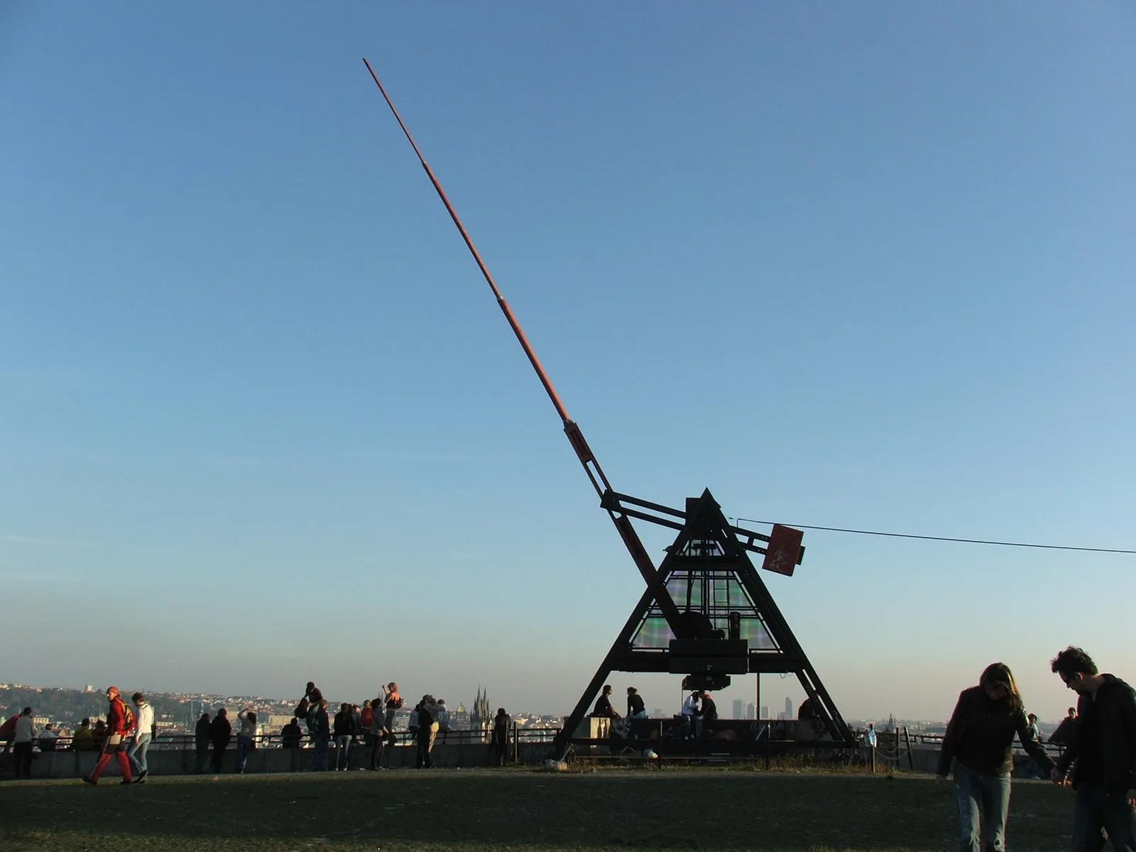 The giant metronome on the Letná terrace in Prague, a long red arm on a dark steel frame silhouetted against the evening sky, with people standing along the parapet above the city