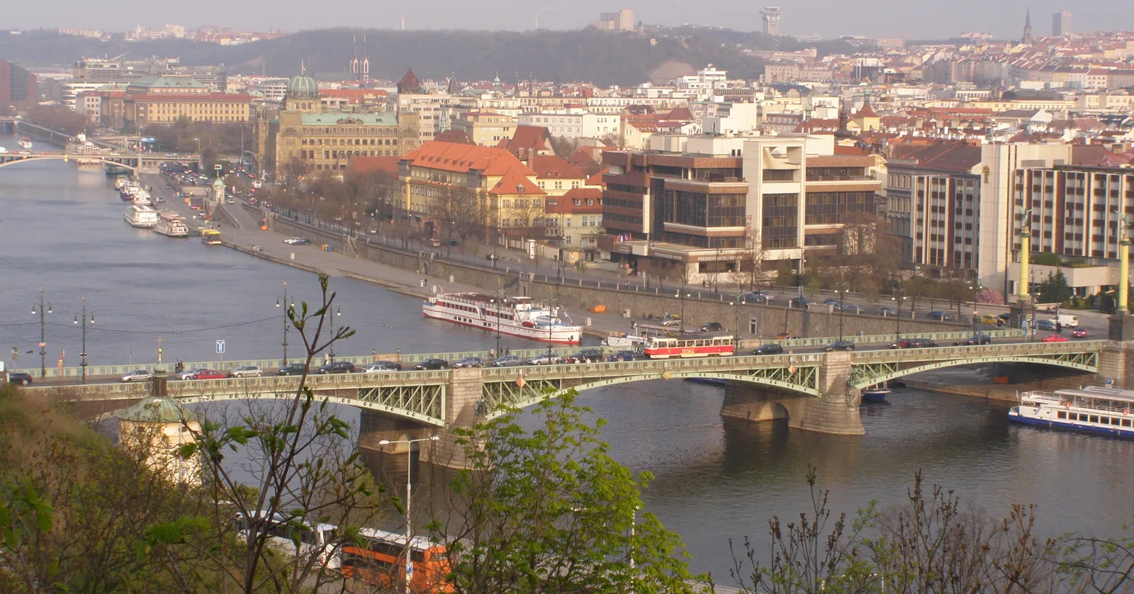 The view south from the Letná bluff in Prague over the green iron Čechův Bridge, with a red tram crossing it, boats moored along the Vltava and the roofs of the Old Town on the far bank