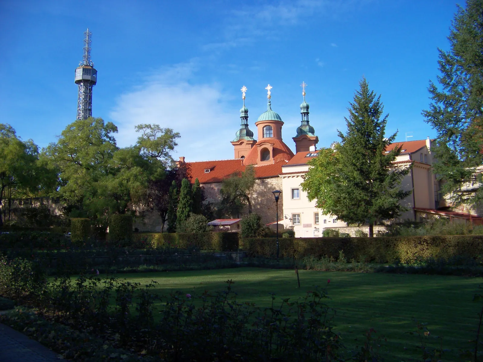 The steel lattice Petřín Lookout Tower rising above trees beside the twin green domes and red roof of the Church of St Lawrence, seen across a lawn in the rose garden