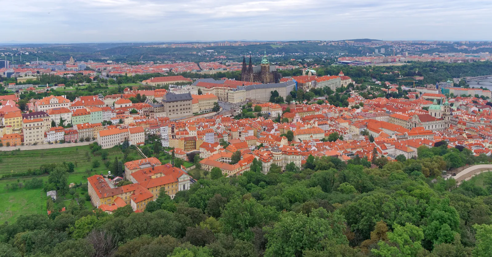 The view north from Petřín Hill over the red-tiled roofs of Malá Strana to Prague Castle and the spires of St Vitus Cathedral, with the green treetops of the Petřín slopes in the foreground