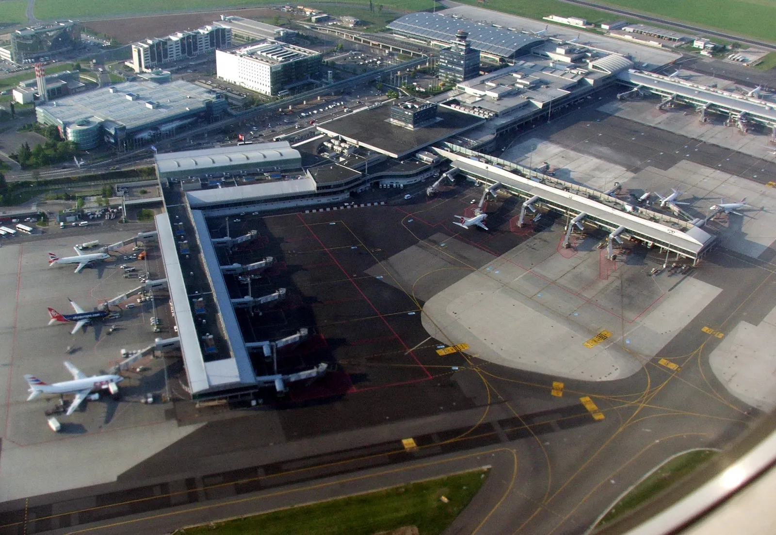 Aerial view of the piers and aprons at Václav Havel Airport Prague, with airliners parked at jet bridges along two finger piers and the control tower behind them