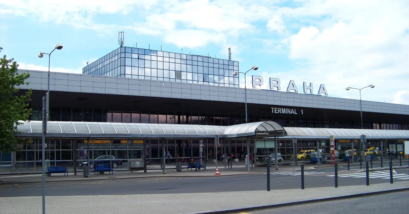 The kerbside frontage of Terminal 1 at Václav Havel Airport Prague, a long low grey building with PRAHA and TERMINAL 1 in large letters across the upper facade and a glass canopy over the pavement below