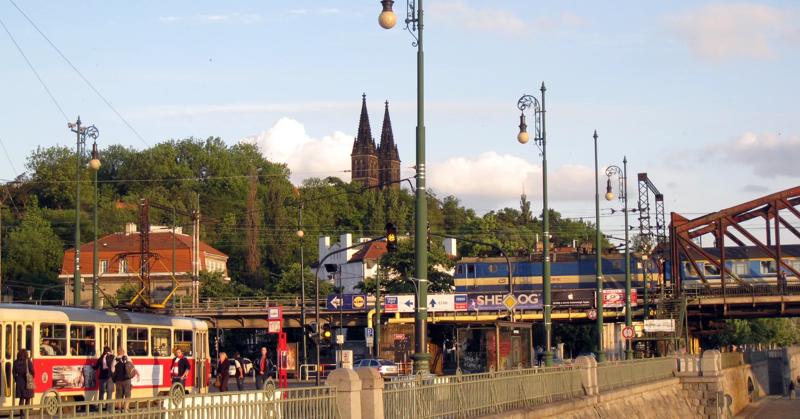 The Vyšehrad rock in Prague seen from the riverside embankment, the twin dark spires of the Basilica of St Peter and Paul rising above wooded slopes, with a tram and the iron railway bridge in the foreground