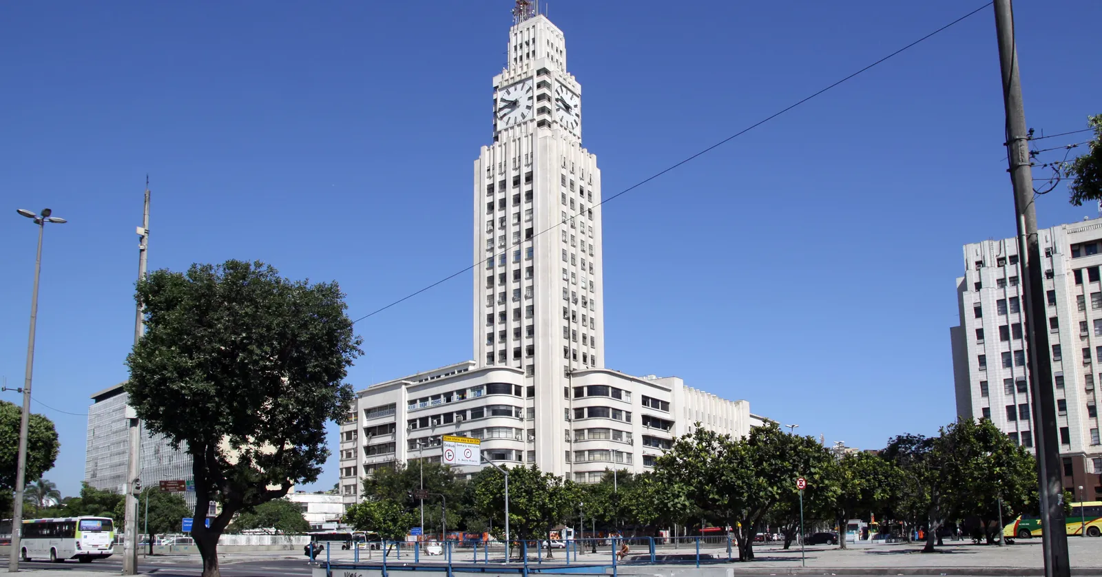 The Central do Brasil station tower in Rio de Janeiro, a stepped white 1940s block with a large clock face near the top, seen across an open square with trees and traffic in front of it under a clear blue sky