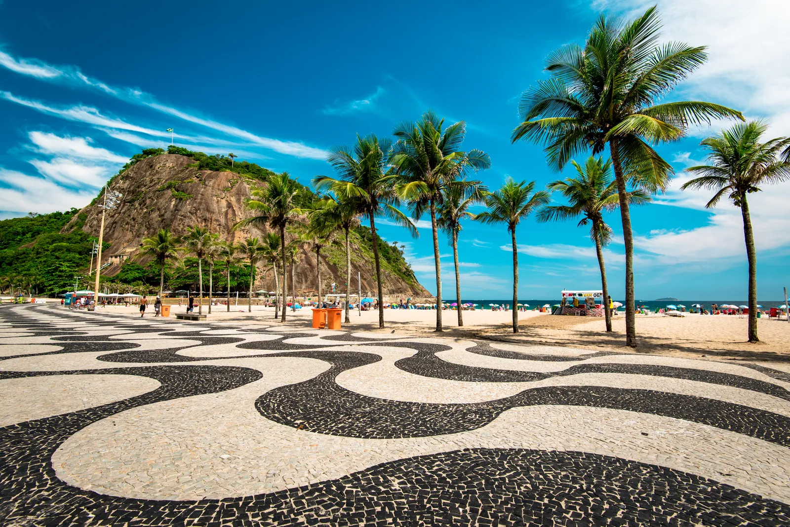 The promenade along Copacabana beach in Rio de Janeiro, paved in a black-and-white wave pattern of small stones, with a row of palms along it, the sand and beach umbrellas beyond and the rocky hill of Leme at the end