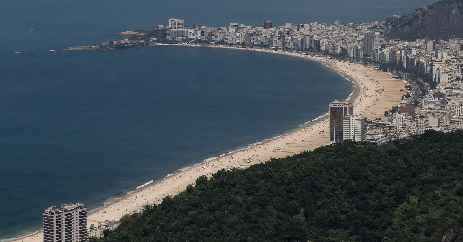 Copacabana beach in Rio de Janeiro seen from a hillside above, the long pale crescent of sand curving away between the sea and a dense wall of apartment towers, with forested slopes in the foreground