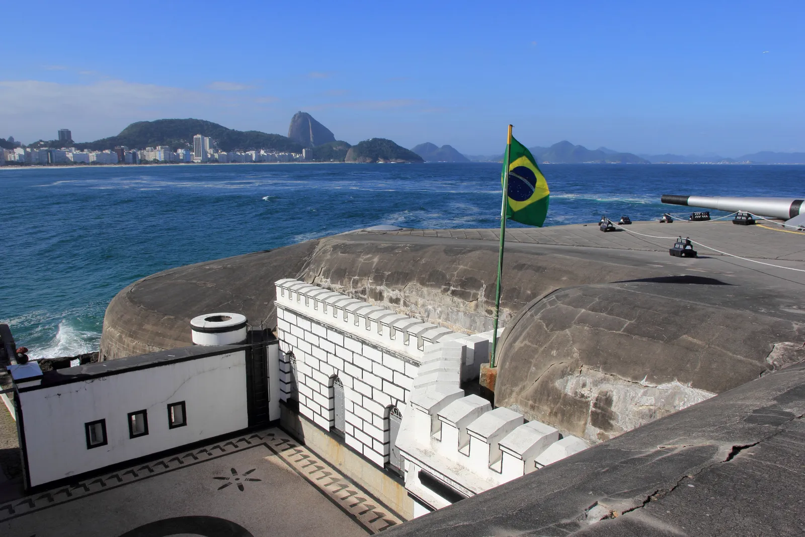 The armoured roof of the Copacabana fort in Rio de Janeiro with a Brazilian flag flying over it and old guns lined up on the terrace beyond, the length of Copacabana beach and Sugarloaf Mountain visible across the water behind