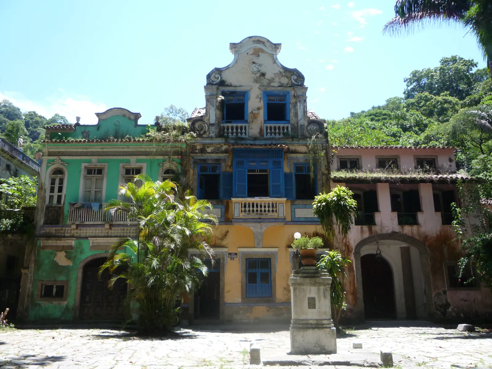 The Largo do Boticário in Rio de Janeiro, a small cobbled square enclosed by green, blue, yellow and pink neocolonial houses with peeling paint and plants growing from the balconies, forest rising behind them