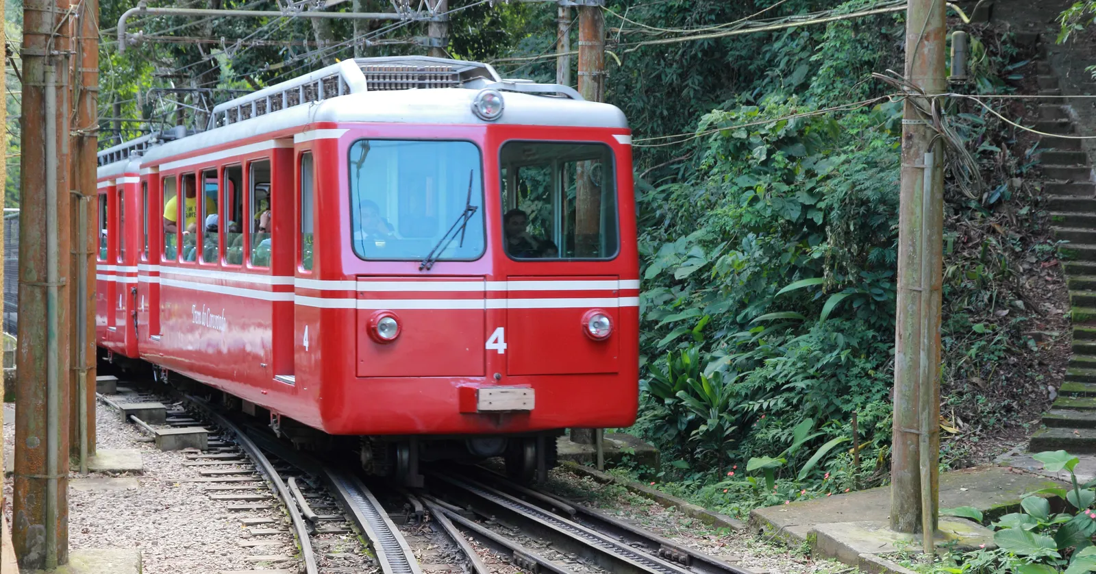 The red Corcovado rack railway train standing on its track in Rio de Janeiro, surrounded on both sides by dense green forest with overhead wires running above the rails