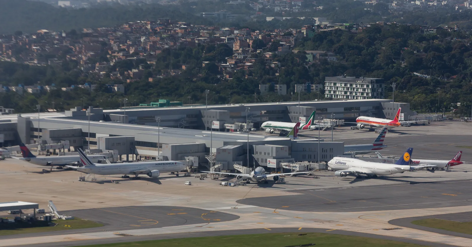 Galeão airport in Rio de Janeiro seen from the air, with a row of airliners at the jet bridges along the low terminal building, service vehicles on the apron and a hillside of houses rising behind