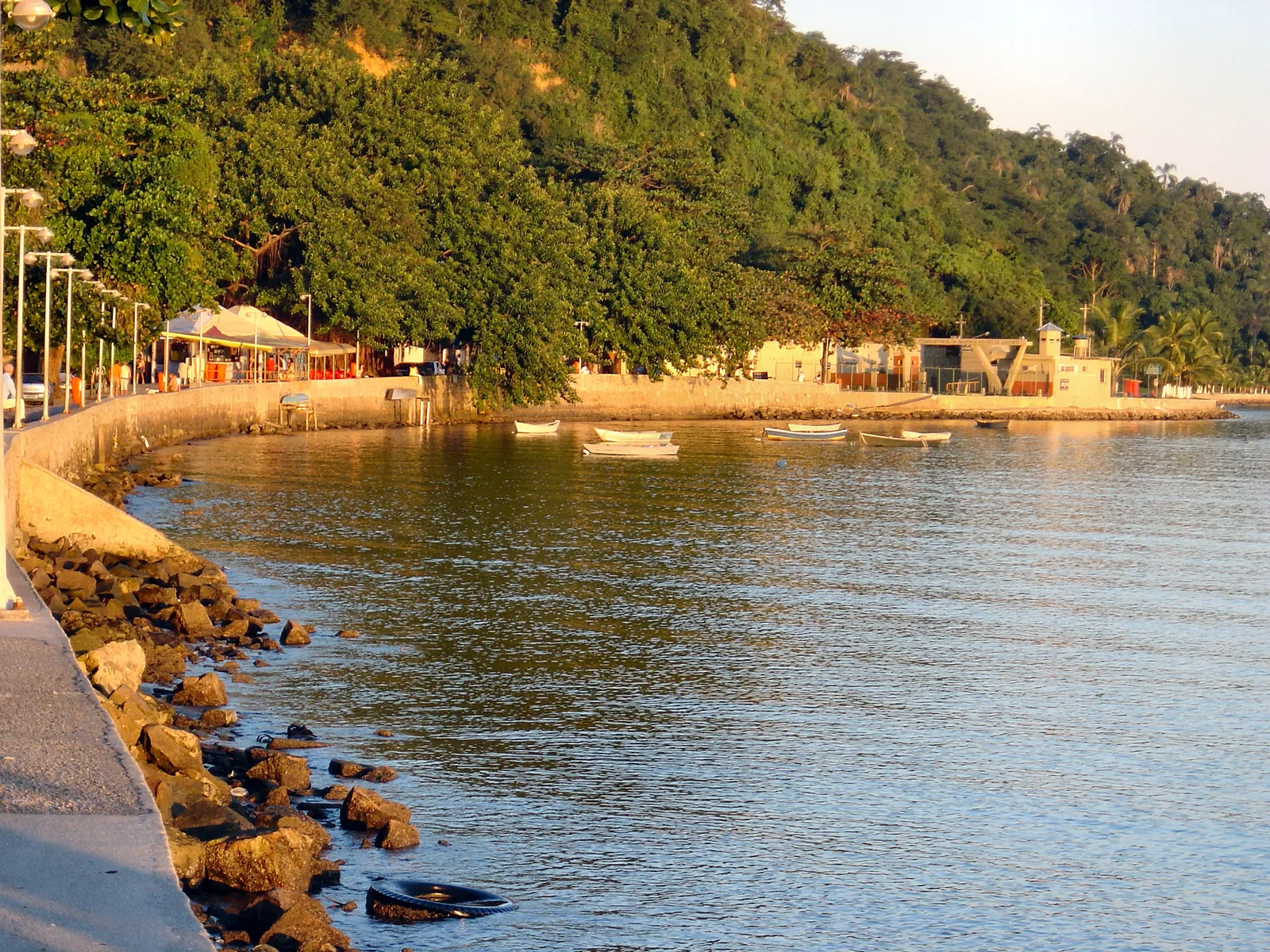 The far end of Praia da Bica on Governador Island in Rio de Janeiro, a curving seawall promenade along calm bay water with small boats moored off a low seafront terrace and a wooded hill rising behind