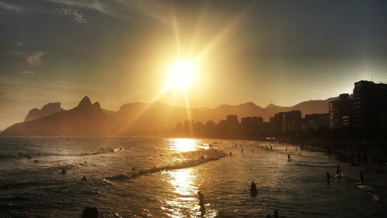 The sun dropping behind the twin peaks of Dois Irmãos at the end of Ipanema beach in Rio de Janeiro, its light spread across the water with swimmers in the surf below