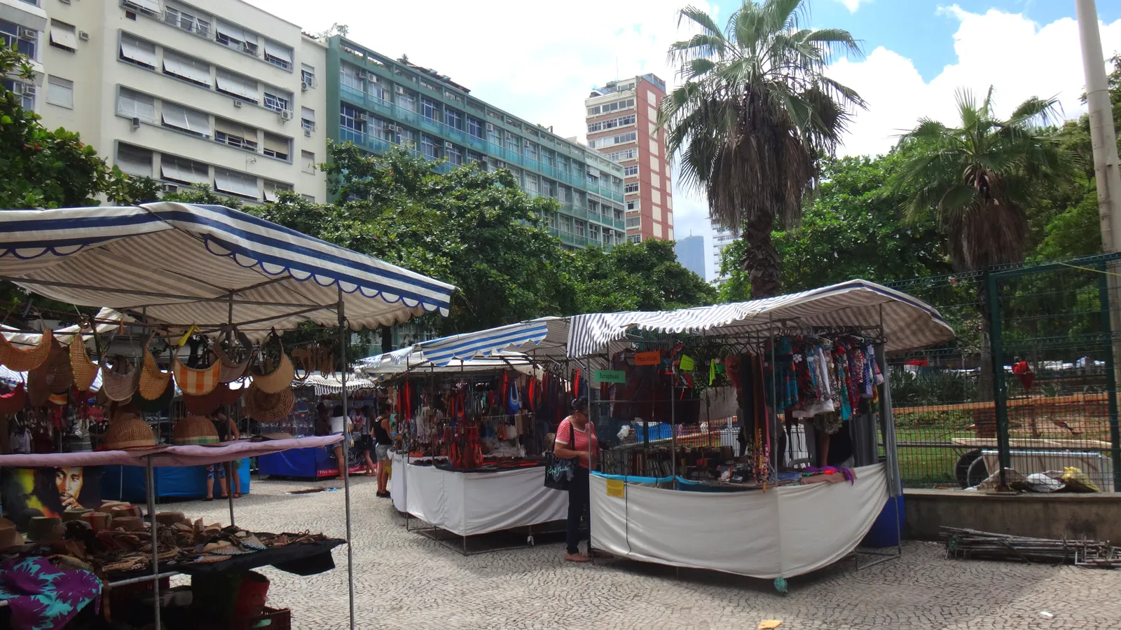 Market stalls under striped awnings on Praça General Osório in Ipanema, Rio de Janeiro, hung with leather bags and clothes, with palms and apartment blocks behind them