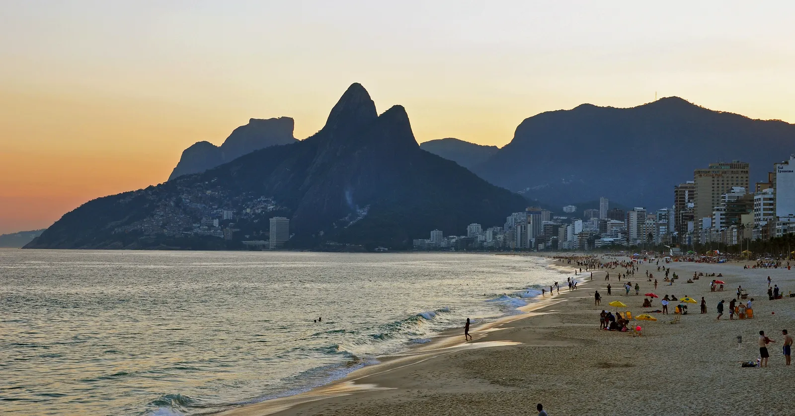Ipanema beach in Rio de Janeiro late in the day, people scattered along the wet sand, apartment blocks running back along the shore on the right and the twin peaks of Dois Irmãos rising dark at the far end