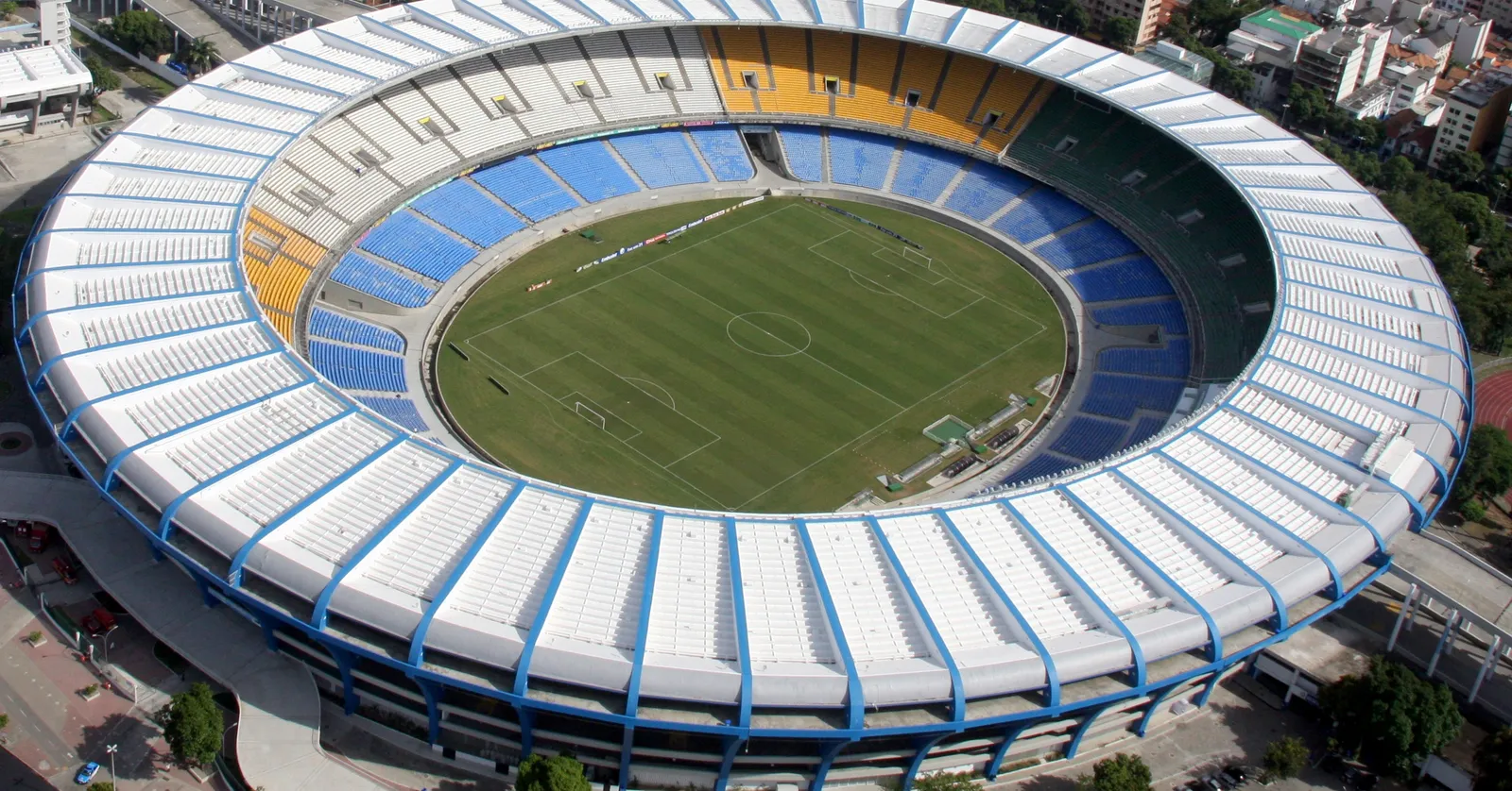 Maracanã stadium in Rio de Janeiro from the air, a single ring of yellow, blue and white seating under a white roof around a green pitch, with the surrounding streets visible at the edges