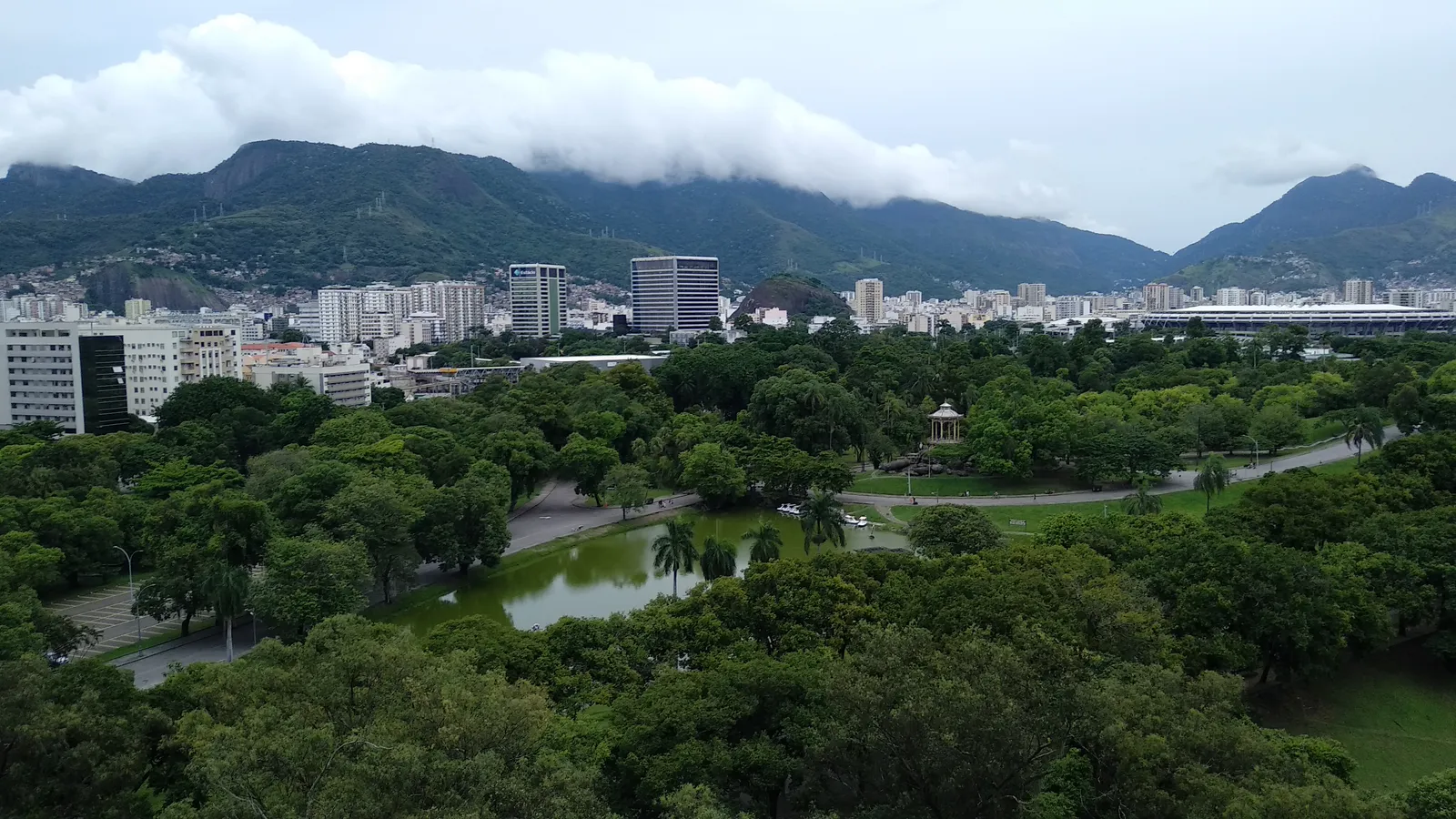 Quinta da Boa Vista in Rio de Janeiro seen from above, a wide green park of old trees around a lake with a small pavilion, city blocks beyond it and cloud sitting on the forested ridge behind