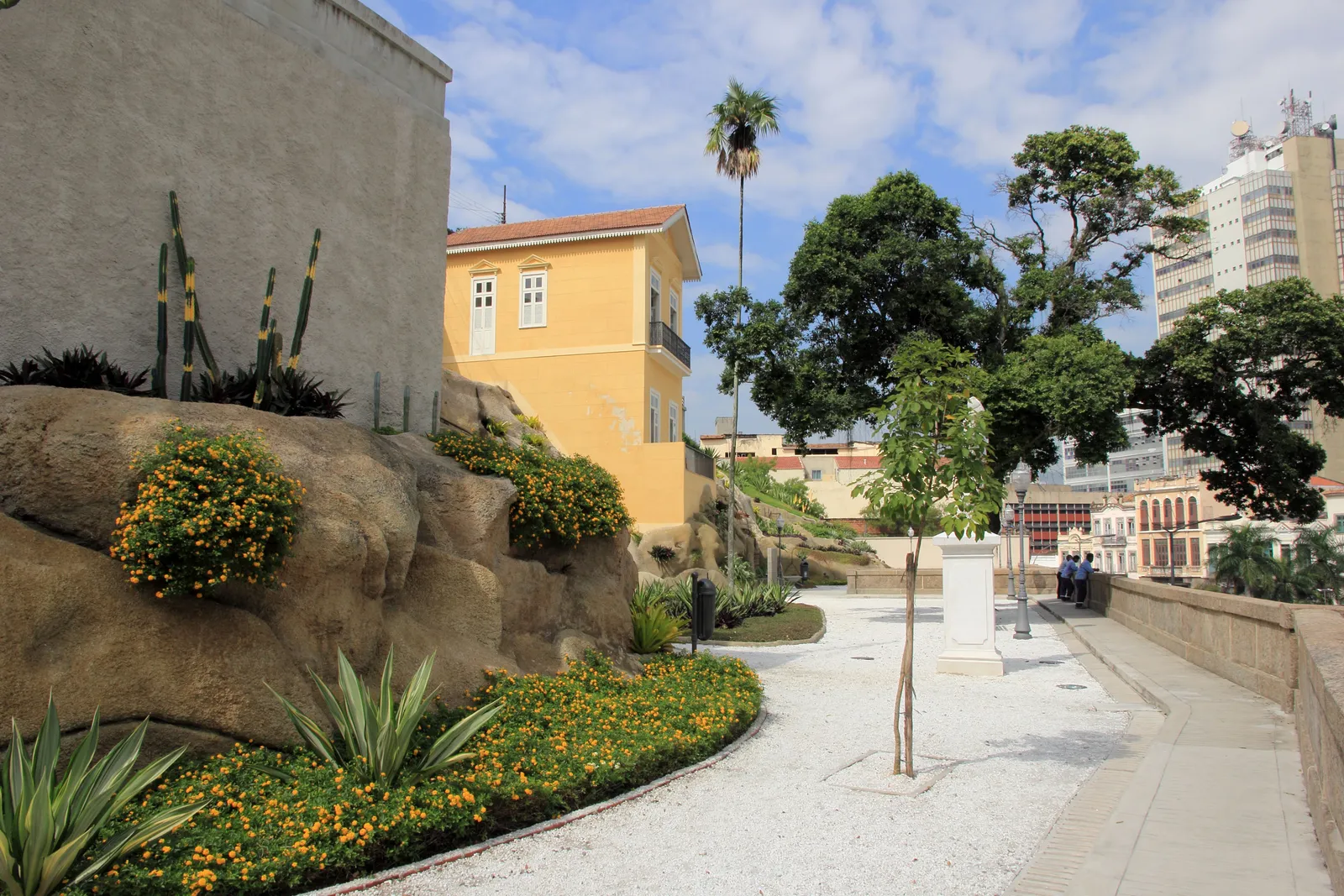 The terraced hanging garden of Valongo in Rio de Janeiro, with flowering beds and palms set among artificial rockwork on a slope, a pale yellow colonial building behind and the port district below