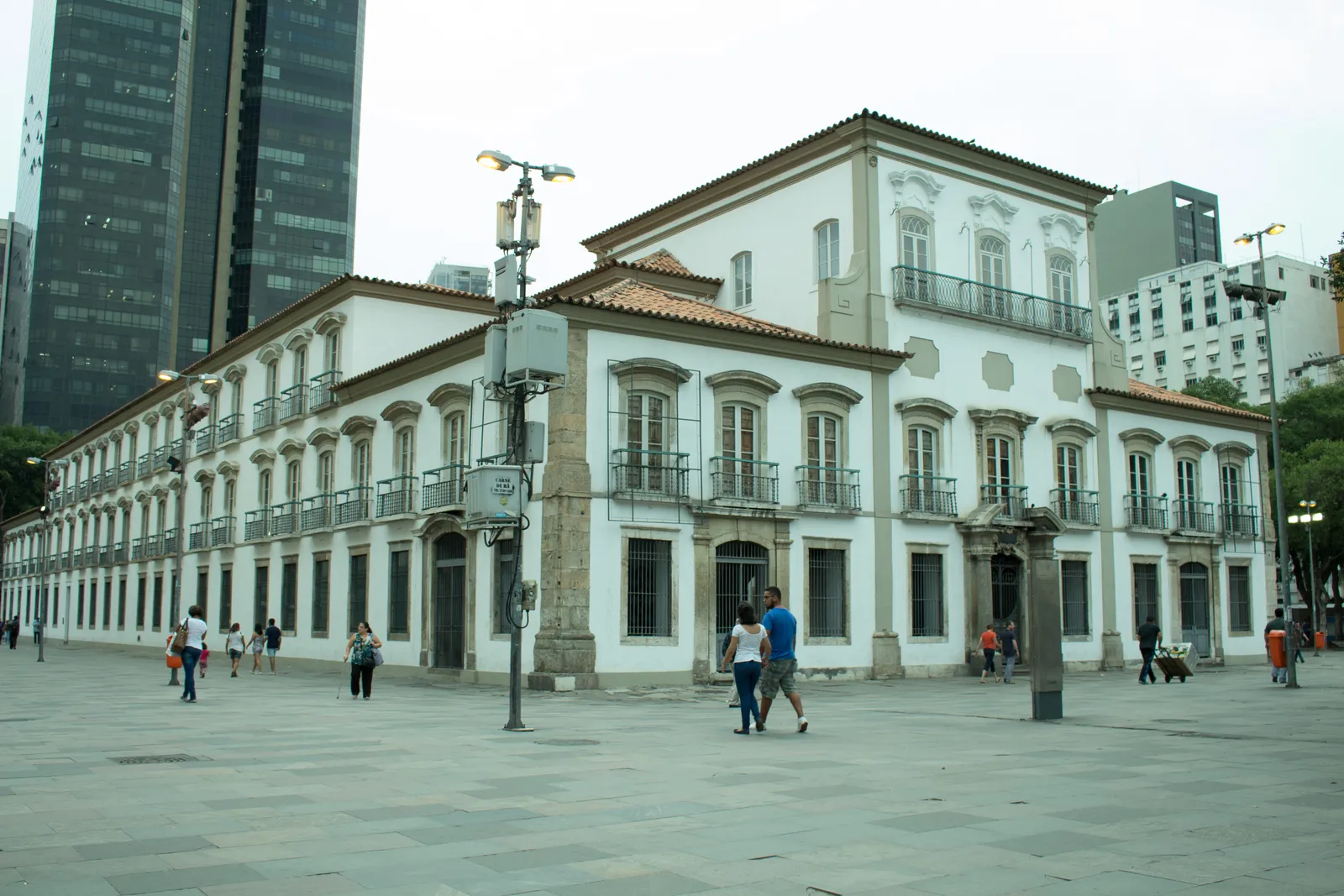 The Paço Imperial on Praça XV in Rio de Janeiro, a long white two-storey colonial palace with a red tiled roof and iron balconies, people crossing the paved square in front of it and office towers behind