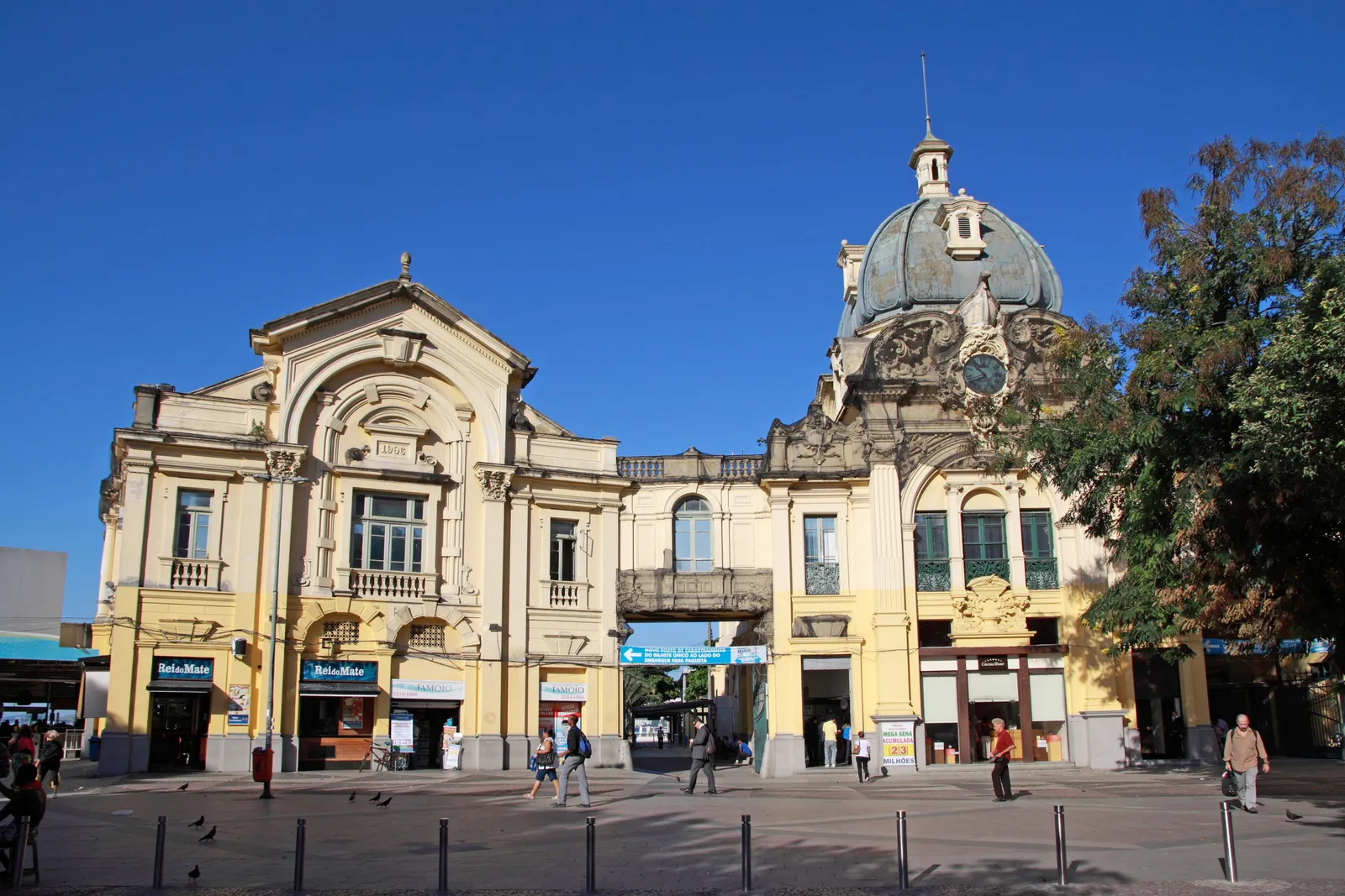 The ferry terminal building on Praça XV in Rio de Janeiro, a pale early 20th-century hall with a green tiled dome and a clock on one corner, people walking across the square in front of it under a clear sky