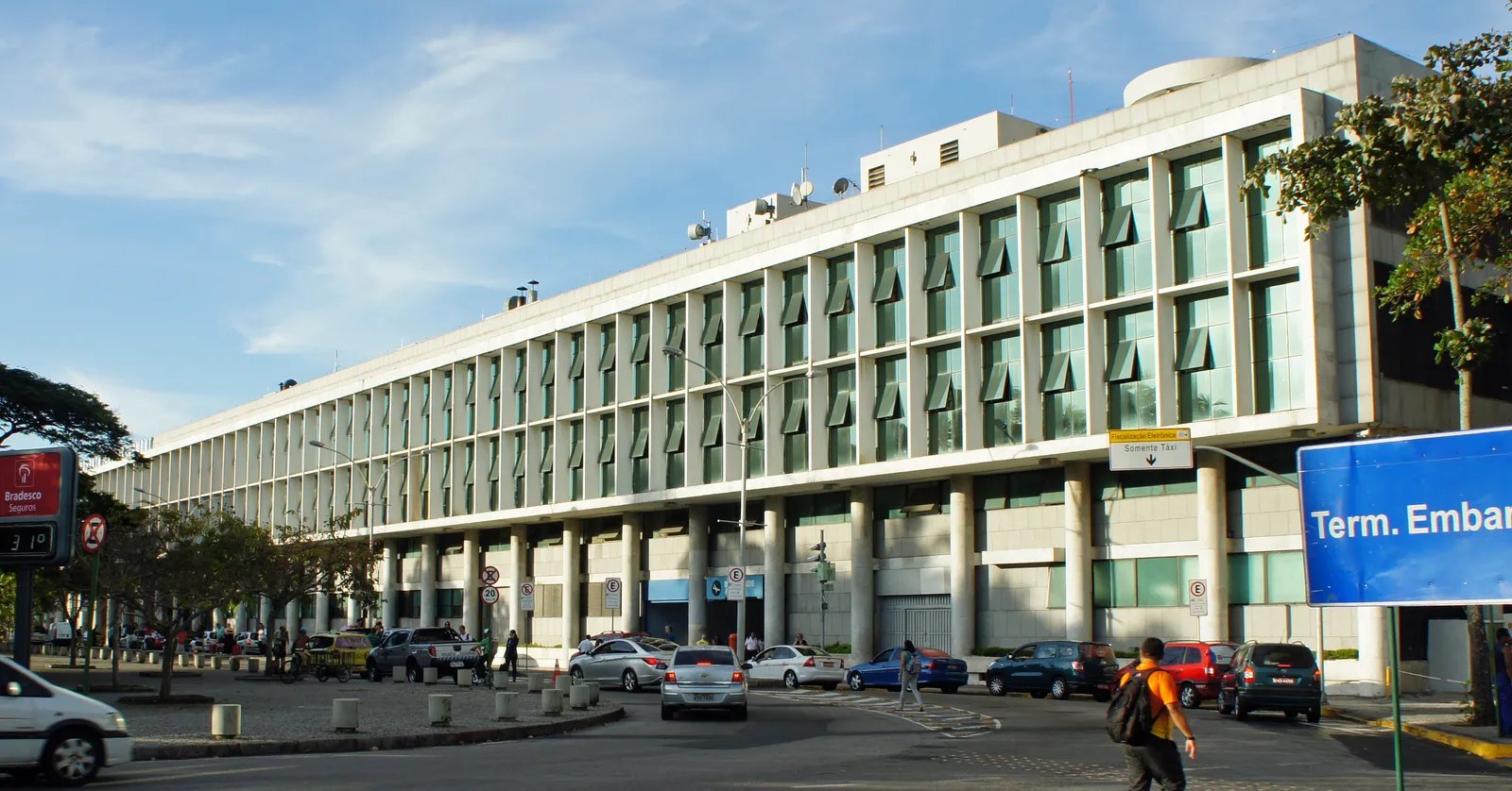 The long pale terminal building at Santos Dumont airport in central Rio de Janeiro, its upper floors set behind a row of square columns, with cars and a taxi rank on the road in front and a blue departures sign at the right