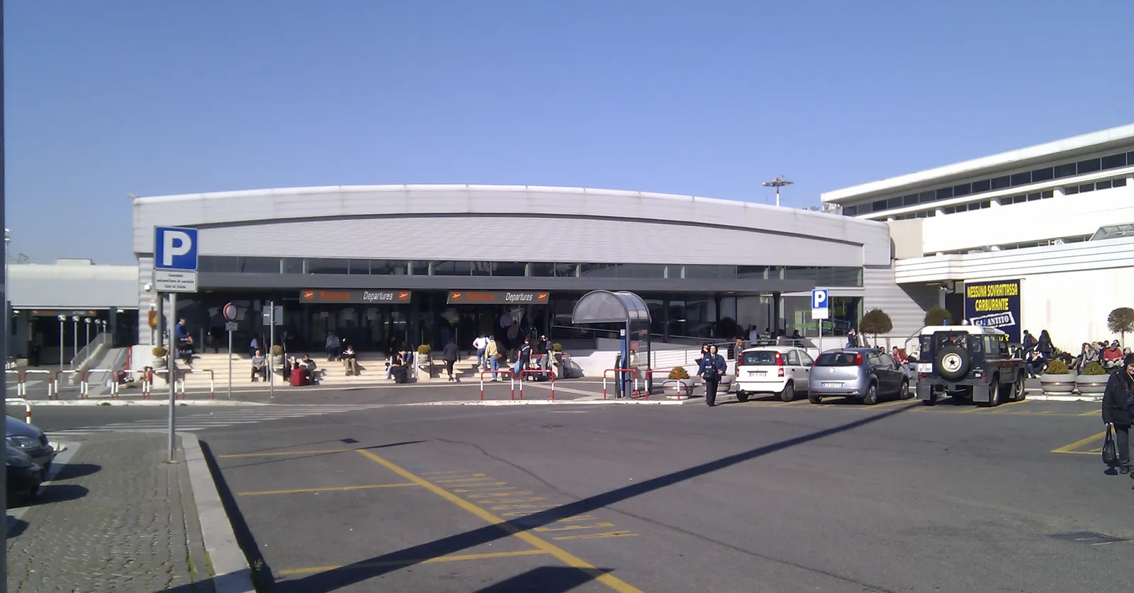 The passenger terminal at Rome Ciampino airport seen from the forecourt, a low building with a curved projecting canopy over its entrance, parked cars and travellers outside under a clear sky