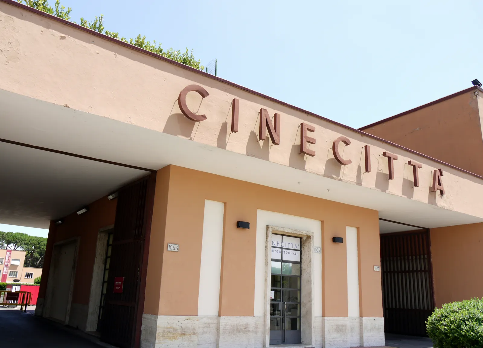 The entrance building of the Cinecitta film studios in Rome, its name spelled out in tall raised letters along the top of a pale ochre facade above the doors