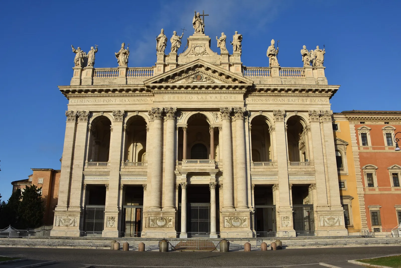 The front of the Basilica of San Giovanni in Laterano in Rome in late afternoon light, a tall columned facade topped by a row of oversized statues standing against a deep blue sky