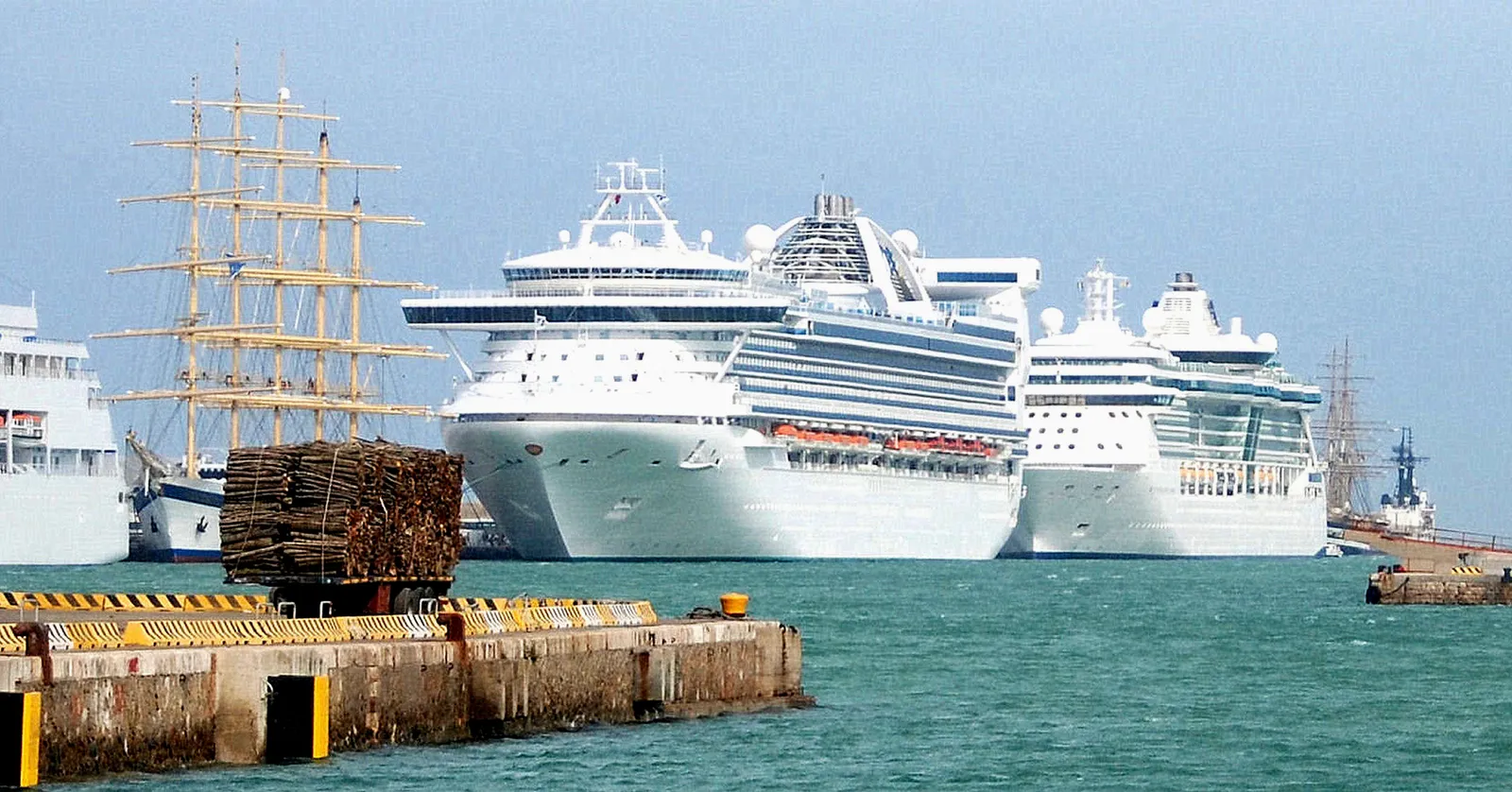 Three large white cruise ships berthed side by side at the port of Civitavecchia near Rome, with the masts of a tall sailing ship to the left
