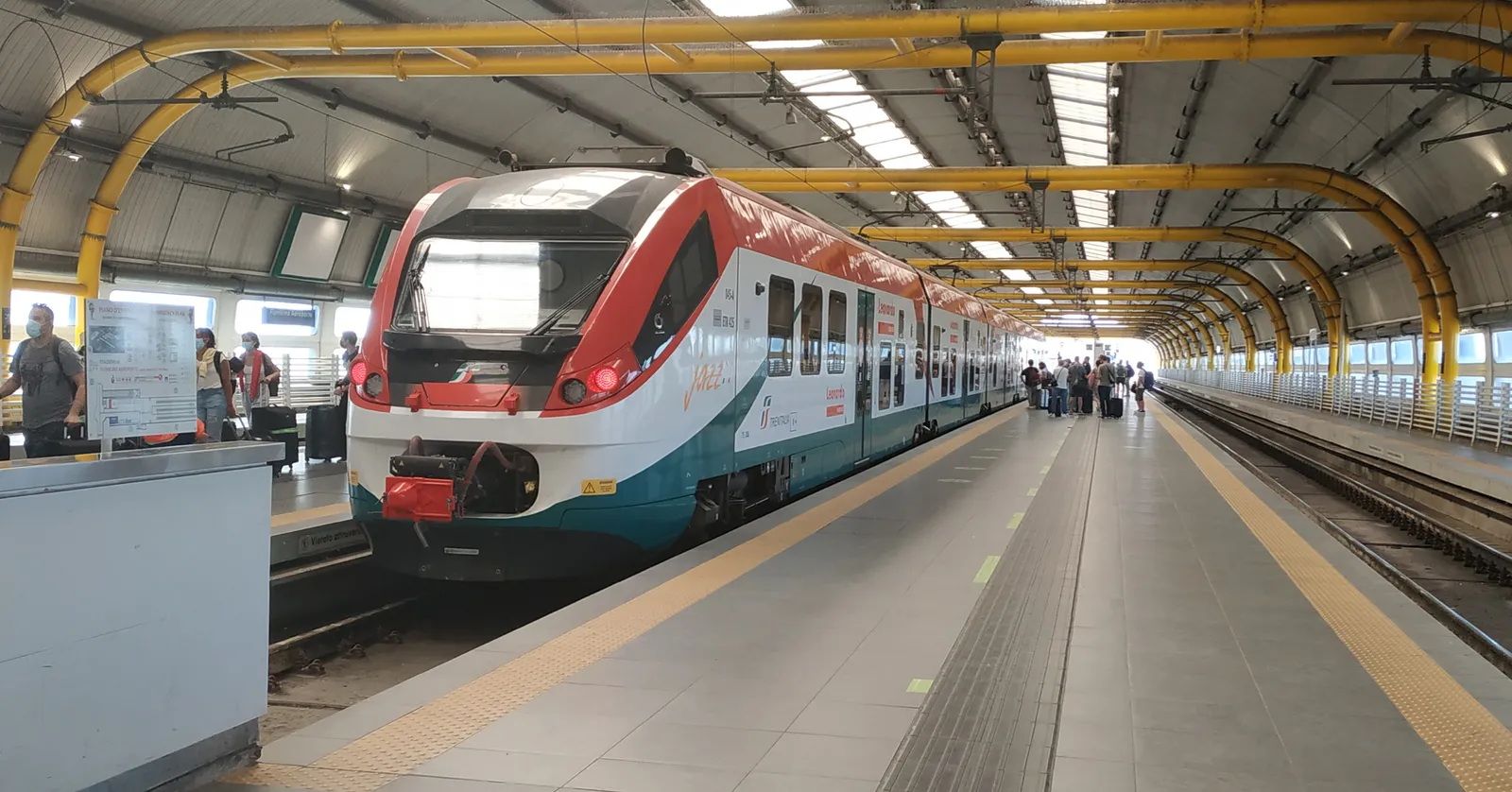 A red, white and green Leonardo Express train at the platform of Fiumicino Aeroporto station in Rome, under a vaulted roof of skylights and yellow steel arches
