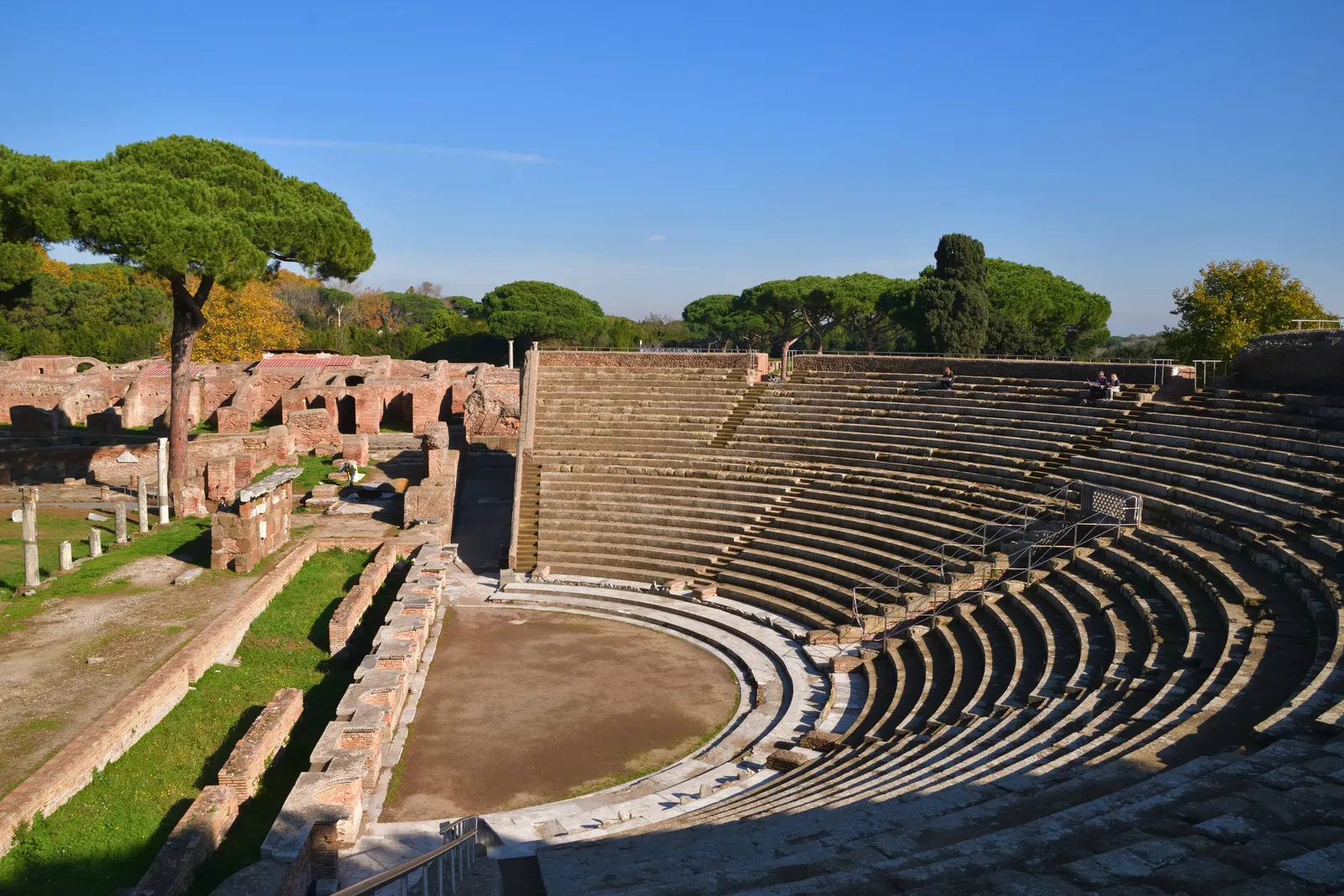 The curved stone seating of the Roman theatre at Ostia Antica near Rome, with umbrella pines and brick ruins behind it under a clear sky