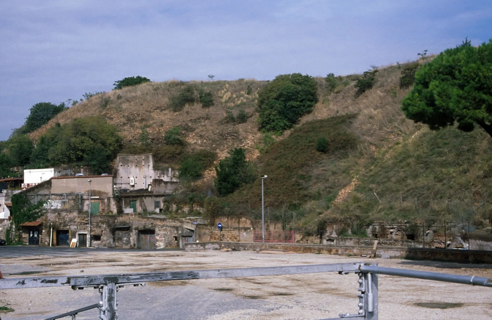 Monte Testaccio in Rome, a low scrub-covered mound rising behind a yard, with small buildings and workshops built into the base of the slope along its foot