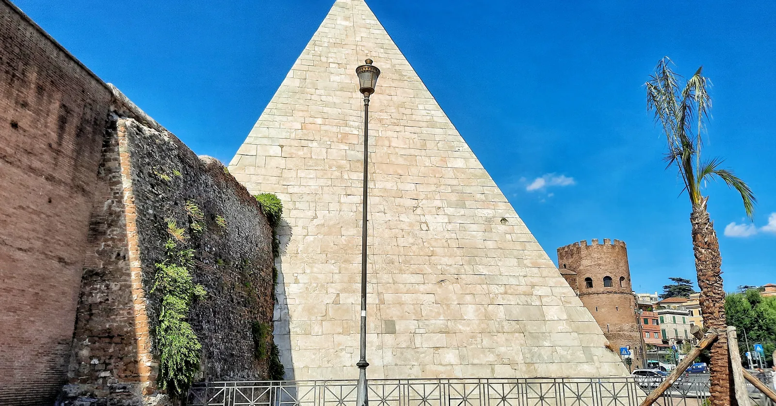 The Pyramid of Cestius in Rome, a steep white marble pyramid rising between a brick section of the Aurelian Walls on one side and a palm tree on the other, under a clear blue sky