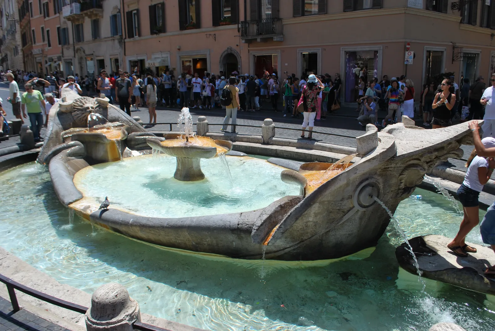 The Fontana della Barcaccia in Rome, a low stone fountain carved as a half-sunk boat with water spilling over its sides, set below street level in the square with people standing around it