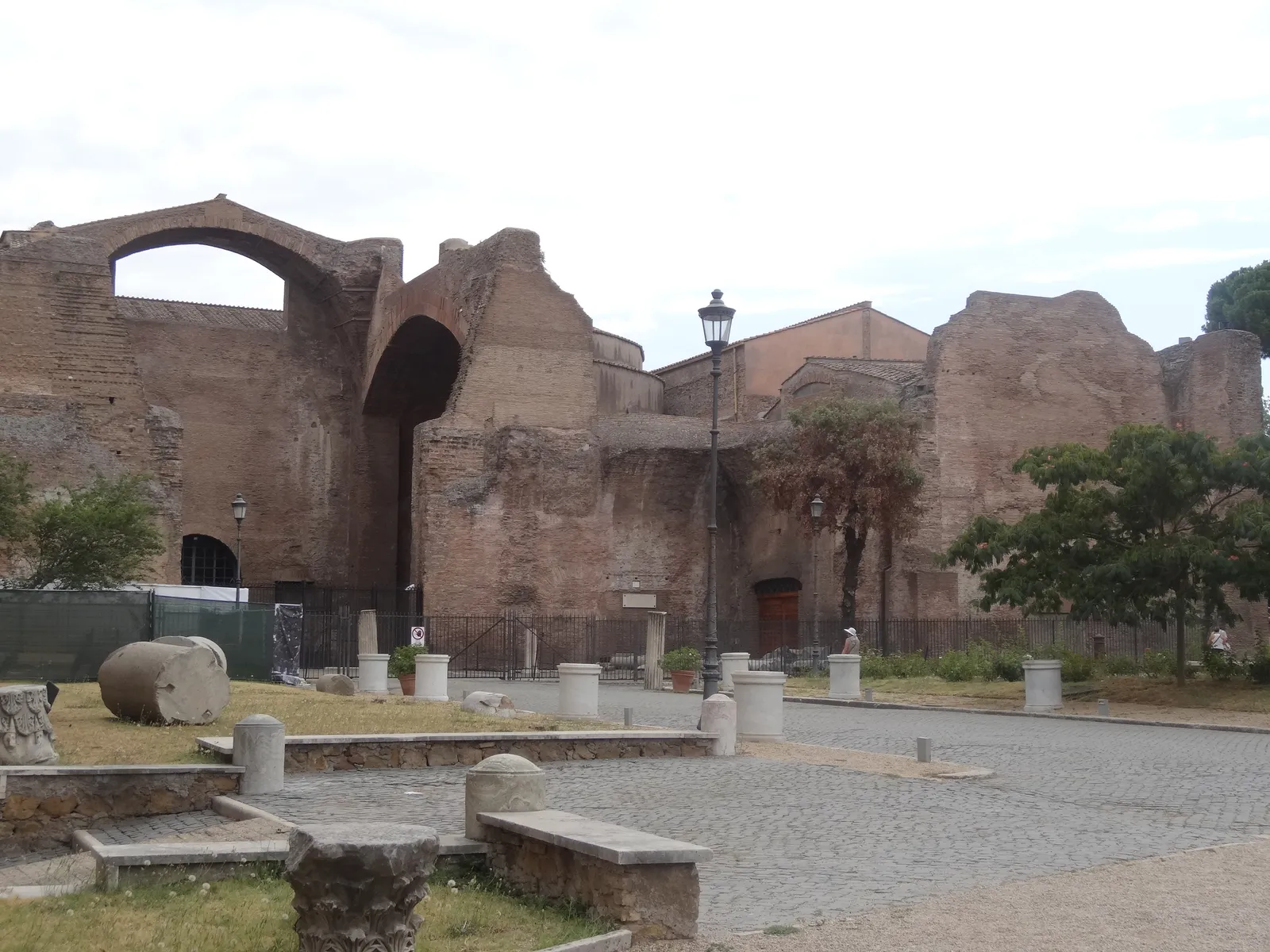 Ruined brick walls and a tall arched vault of the Baths of Diocletian in Rome, with column drums and fragments of stone standing on the gravel forecourt in front of them