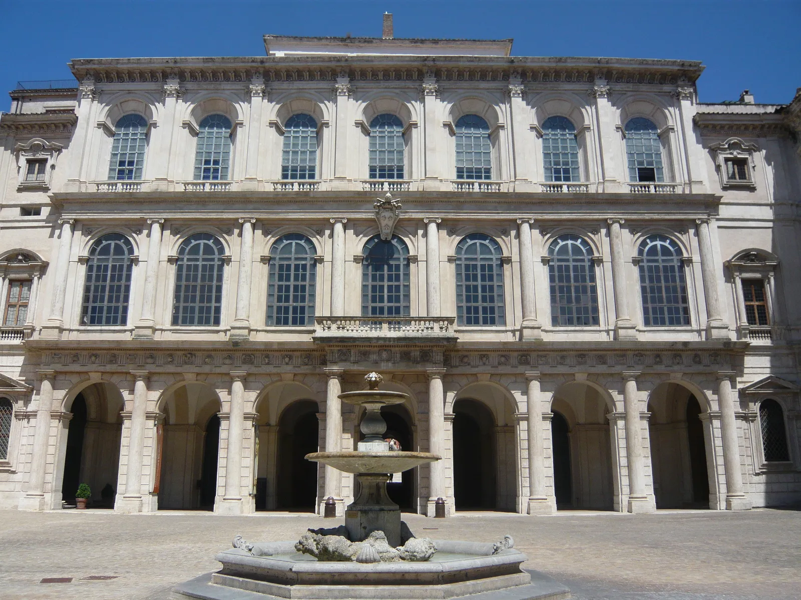 The front of Palazzo Barberini in Rome, a wide pale stone facade with three tiers of arched windows above an open arcaded loggia, and a low fountain standing in the forecourt