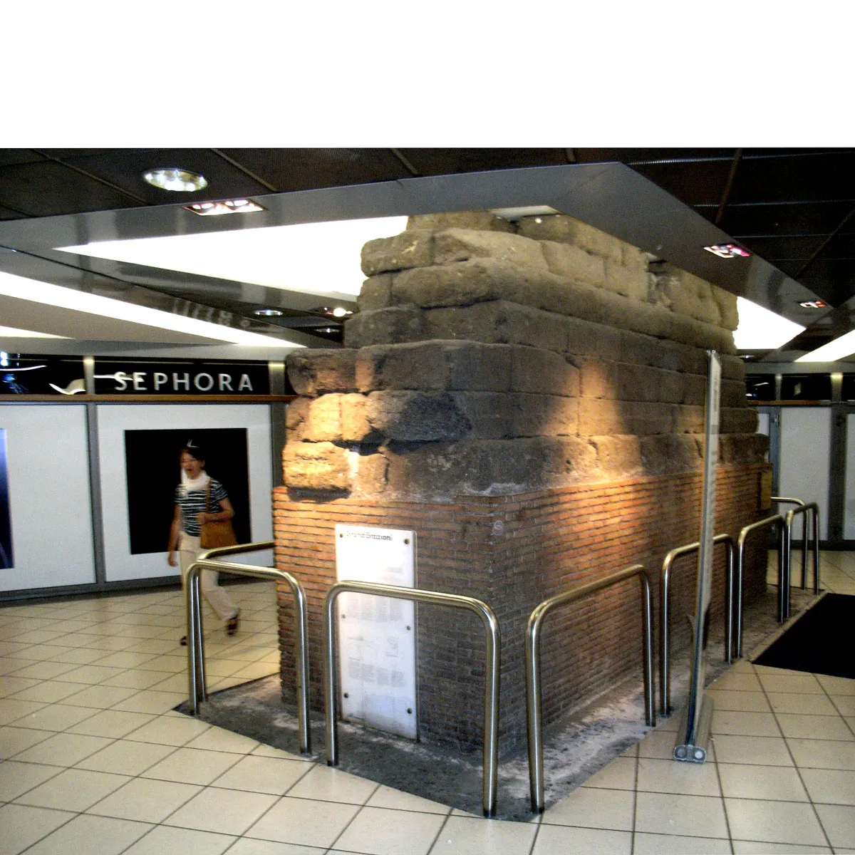 A block of the ancient Servian Wall standing railed off in the middle of the lower-level shopping concourse at Roma Termini, tiled floor and lit shopfronts around it