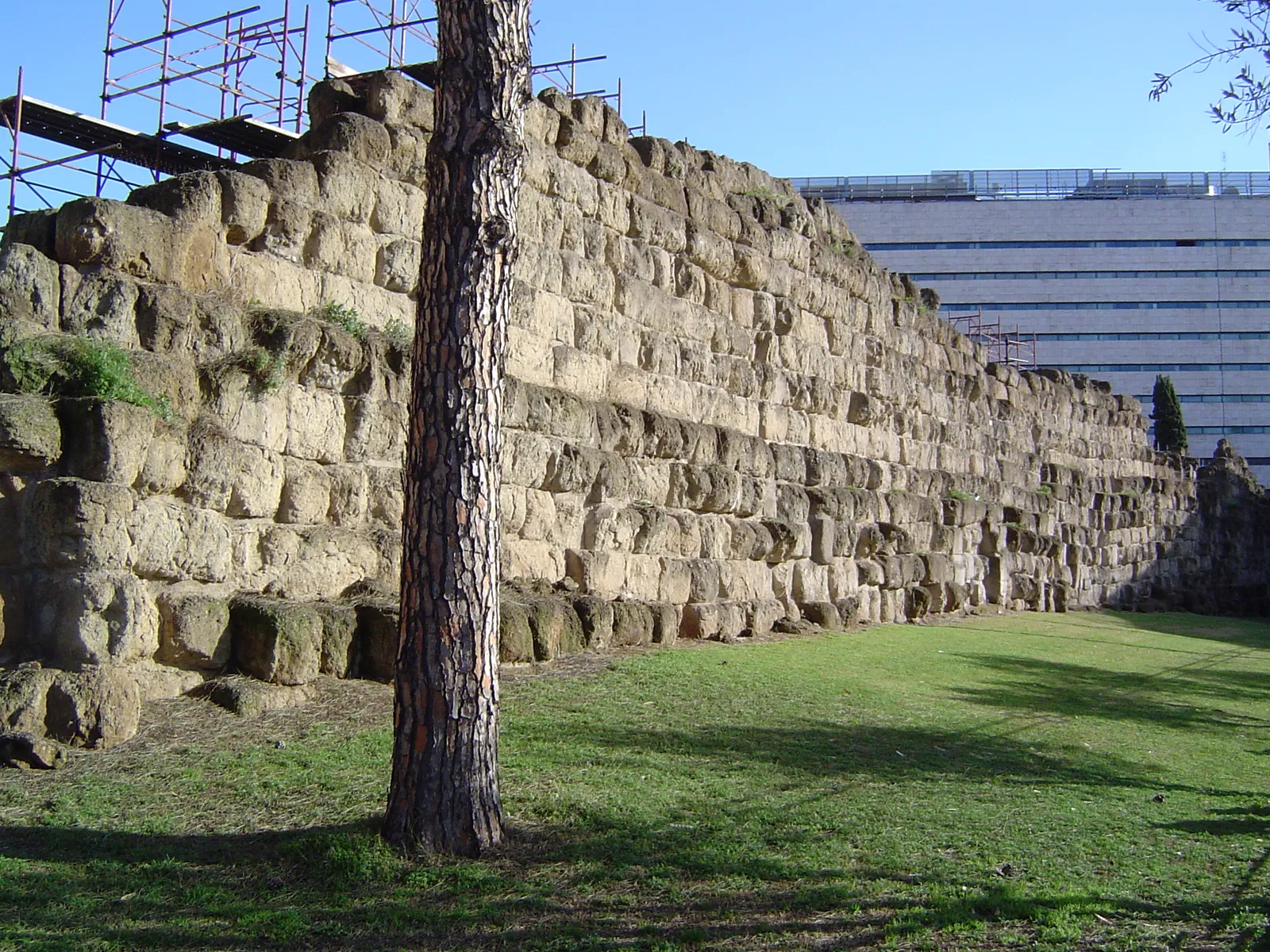 A long stretch of the Servian Wall beside Roma Termini station, courses of squared golden tufa blocks running away across the grass with the station's modern building rising behind it