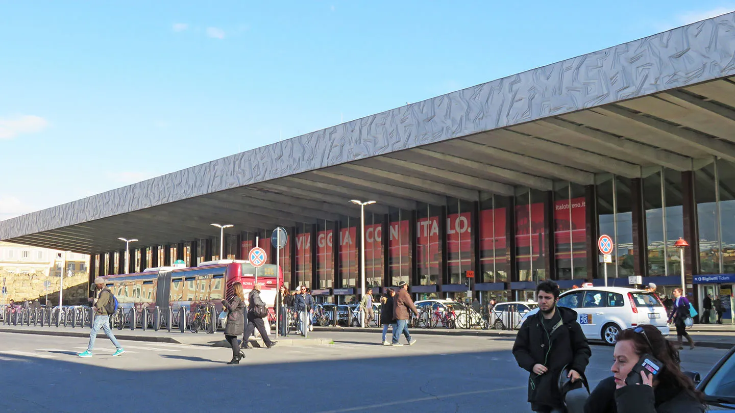 The front of Roma Termini station from Piazza dei Cinquecento, its long cantilevered concrete canopy projecting over the entrance with a glass wall beneath it and buses drawn up outside