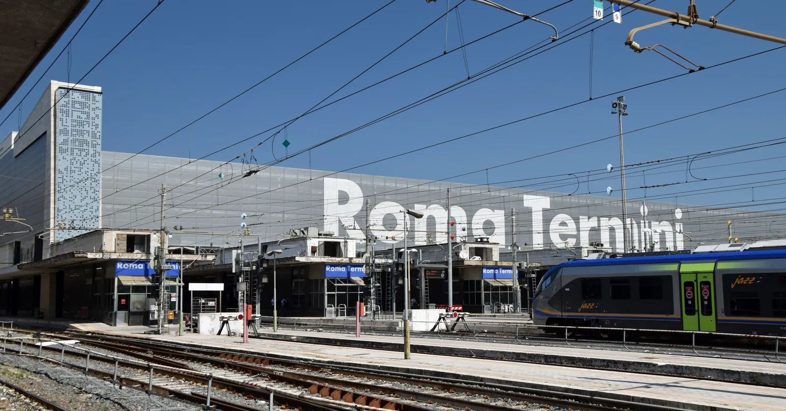 Roma Termini station seen across its tracks, the words ROMA TERMINI in large white letters on the glass end wall of the train shed, with a regional train standing at a platform under overhead wires
