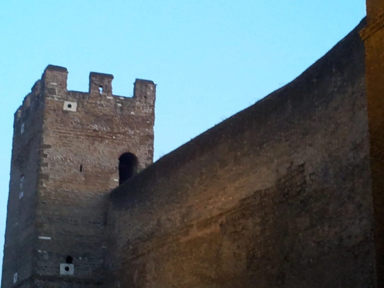 A brick stretch of the Aurelian Walls beside the San Lorenzo quarter in Rome, a square crenellated tower with an arched opening standing at the end of the curtain wall against a clear sky