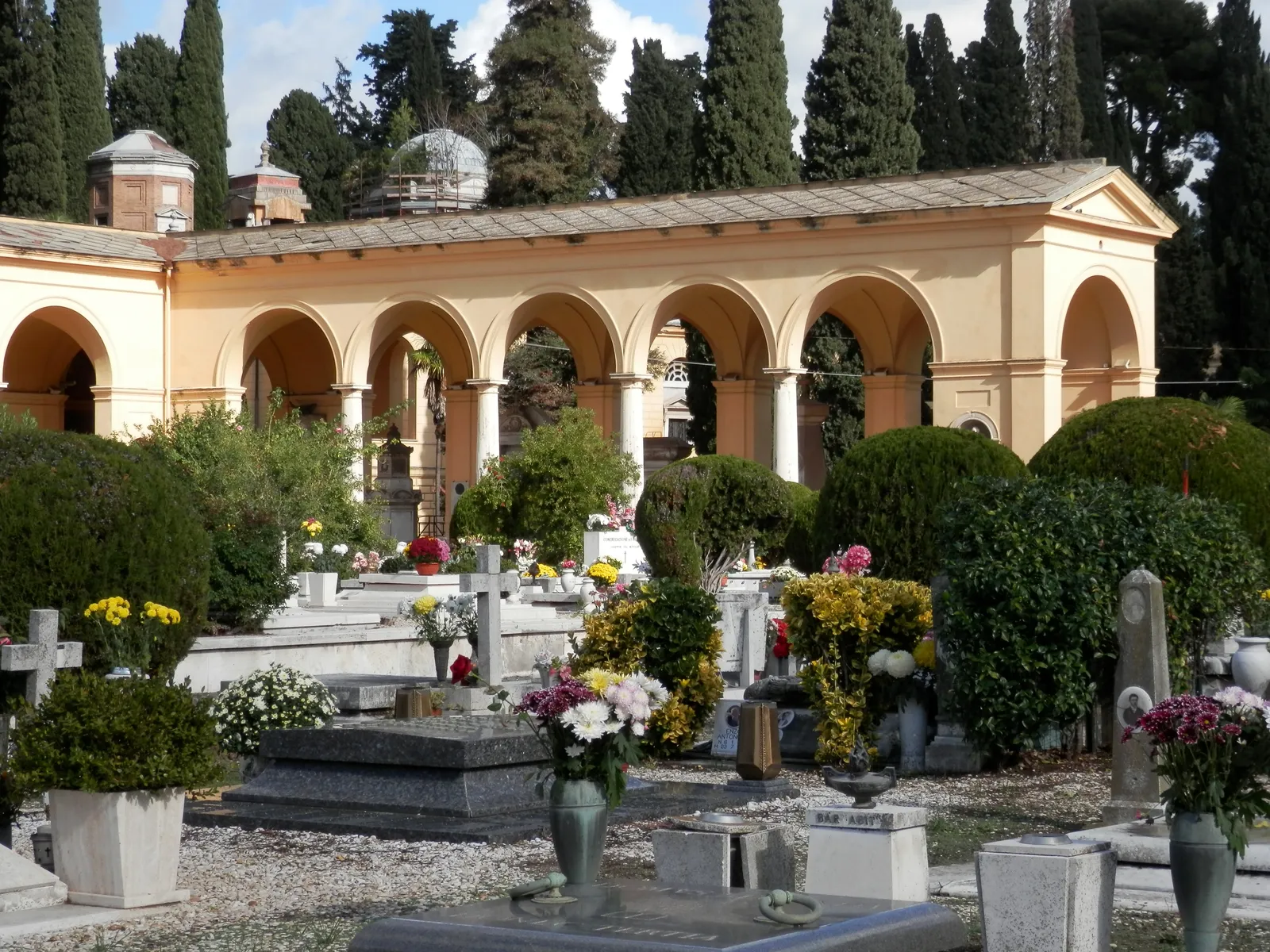 An arcaded gallery of tombs at the Campo Verano cemetery in Rome, ochre arches running along a terrace above rows of graves banked with fresh flowers, cypresses rising behind