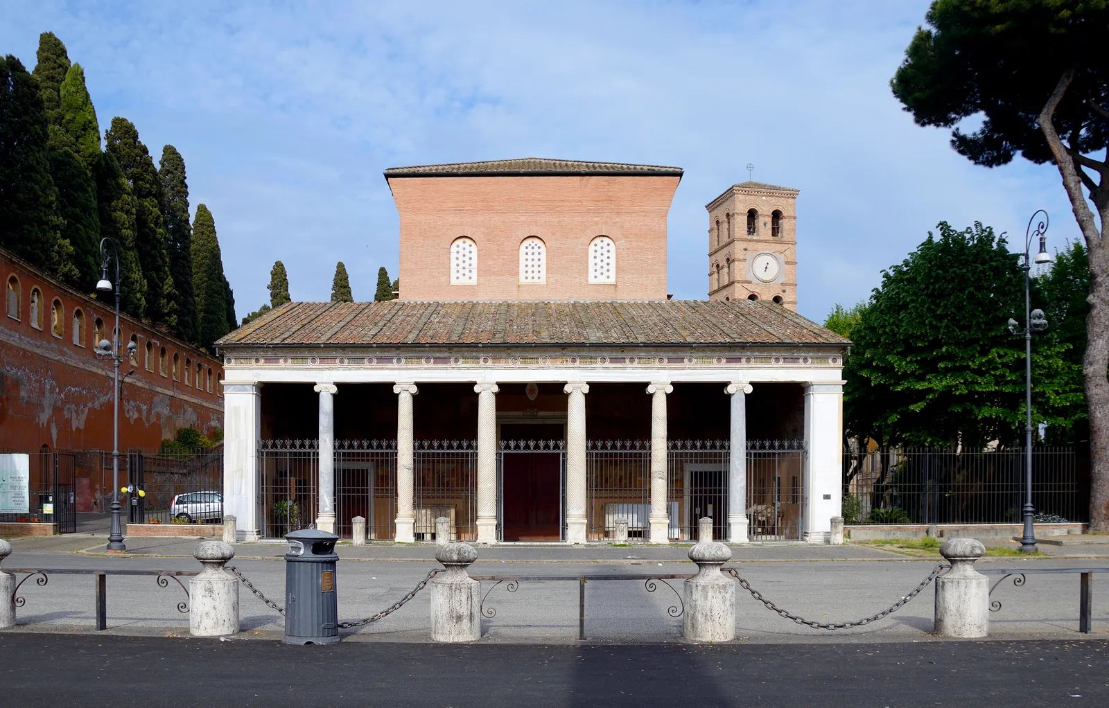 The basilica of San Lorenzo fuori le Mura in Rome, a low columned portico across the front of a plain brick church with a Romanesque bell tower beside it and cypresses behind