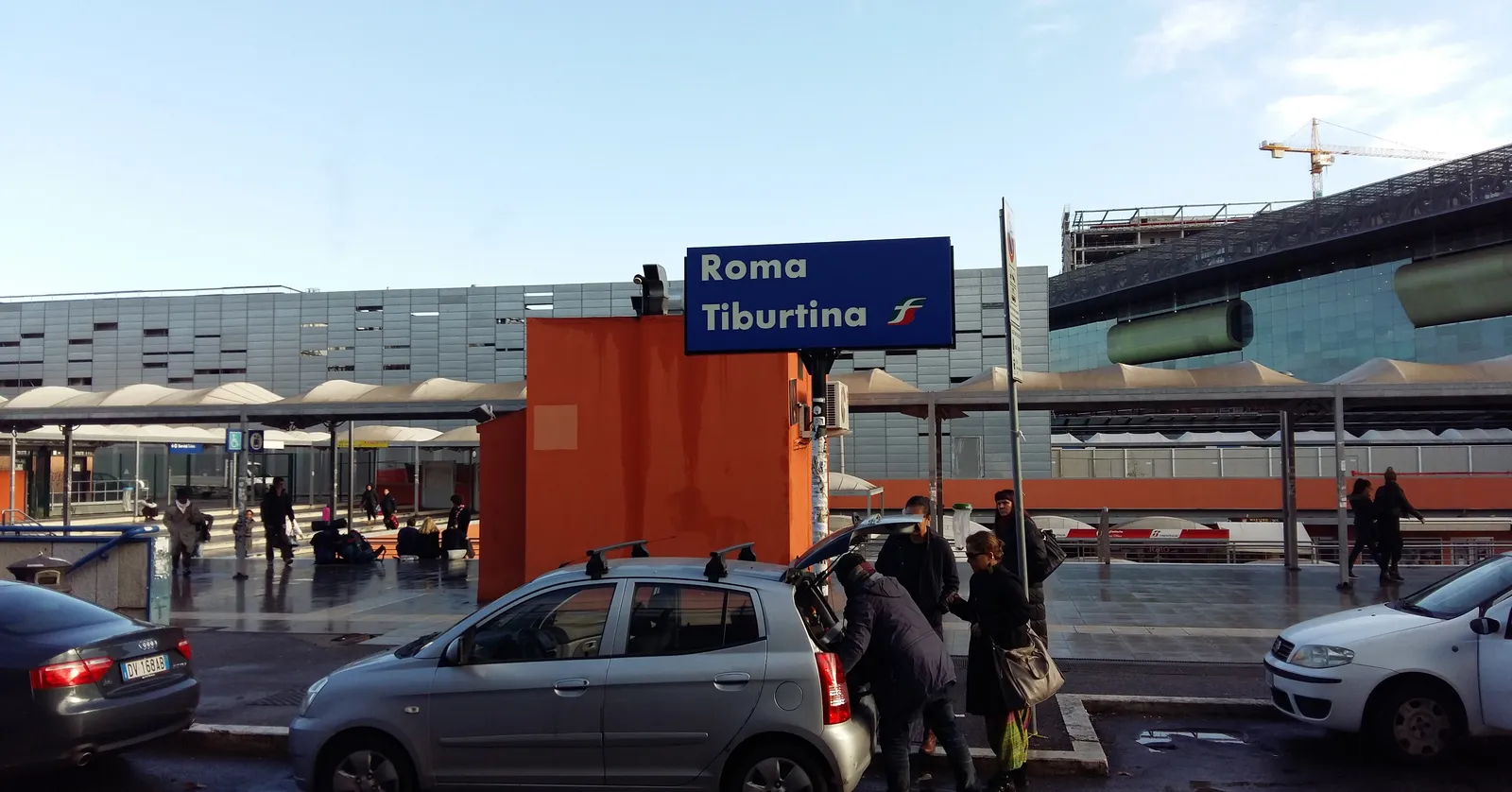 The entrance forecourt at Roma Tiburtina station, a large blue sign reading ROMA TIBURTINA above an orange-panelled facade, with canopies, taxis and travellers crossing in front of it