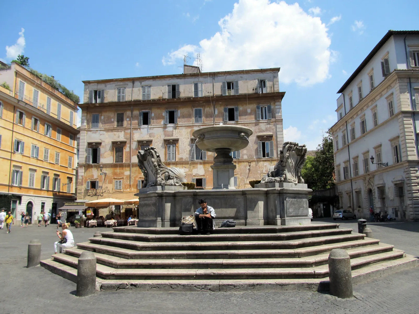 The octagonal fountain in Piazza di Santa Maria in Trastevere in Rome, its stone basin raised on wide steps where people sit, with ochre and cream palazzo fronts closing the square behind